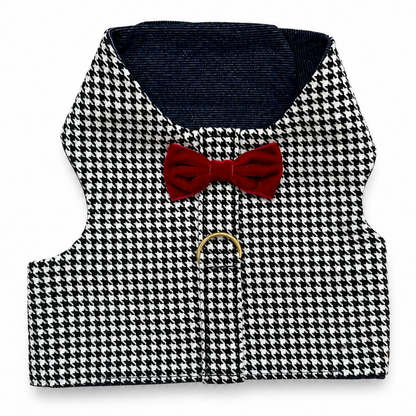 A small black and white houndstooth harness with a dark navy blue lining, a red bow tie, and a gold metal ring on a white background.