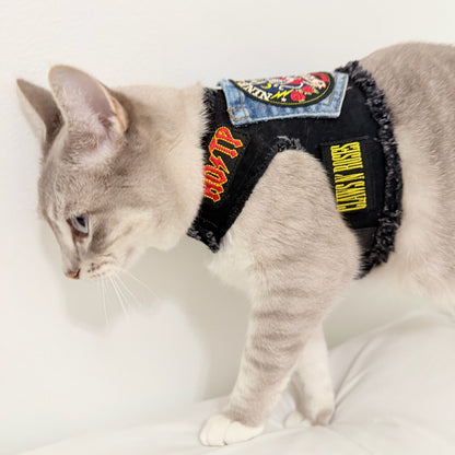 A gray cat wearing a denim and black patchwork pet harness with embroidered patches, standing on a white surface against a white wall.