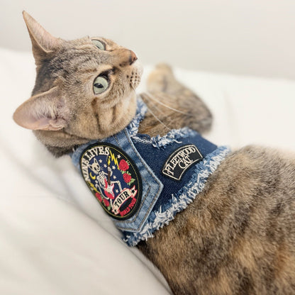 A tabby cat wearing a denim-style pet harness with embroidered patches, lying on a white surface in a softly lit indoor setting.