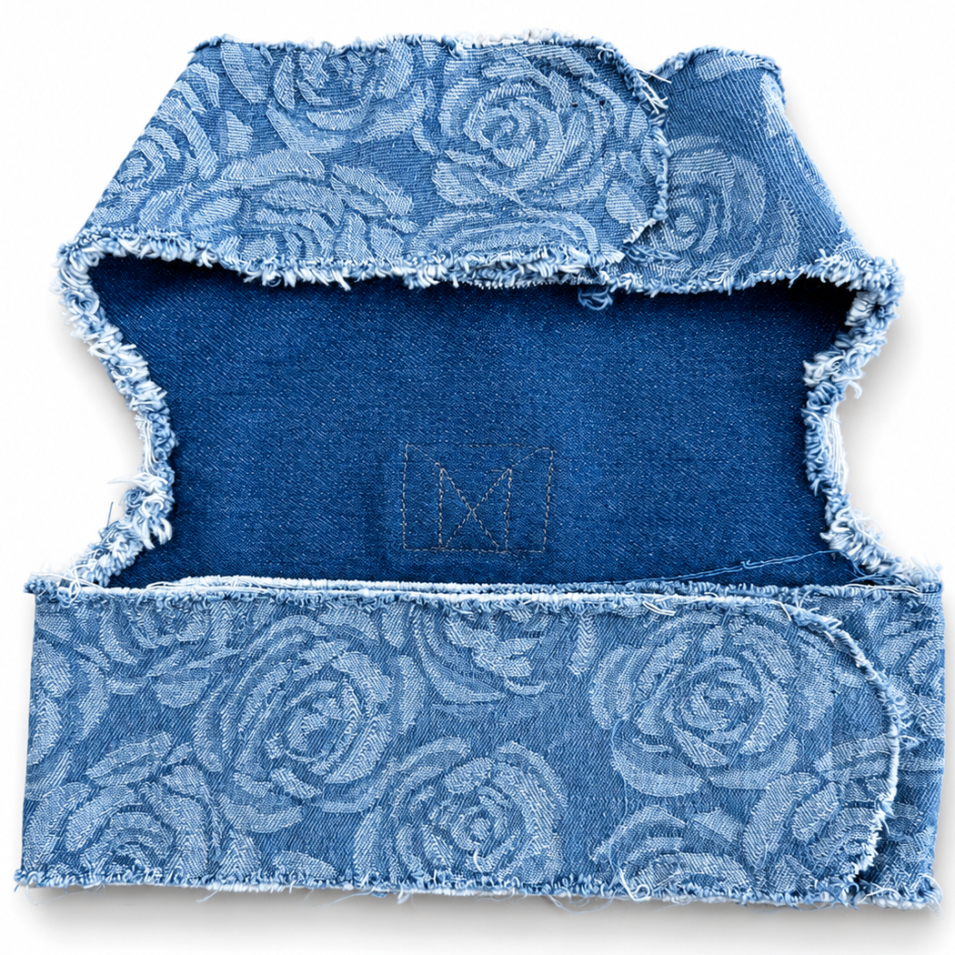 A blue denim harness folded to show the back side, featuring a lighter blue floral rose pattern on the outer fabric and a solid medium blue inner lining. The edges are frayed.