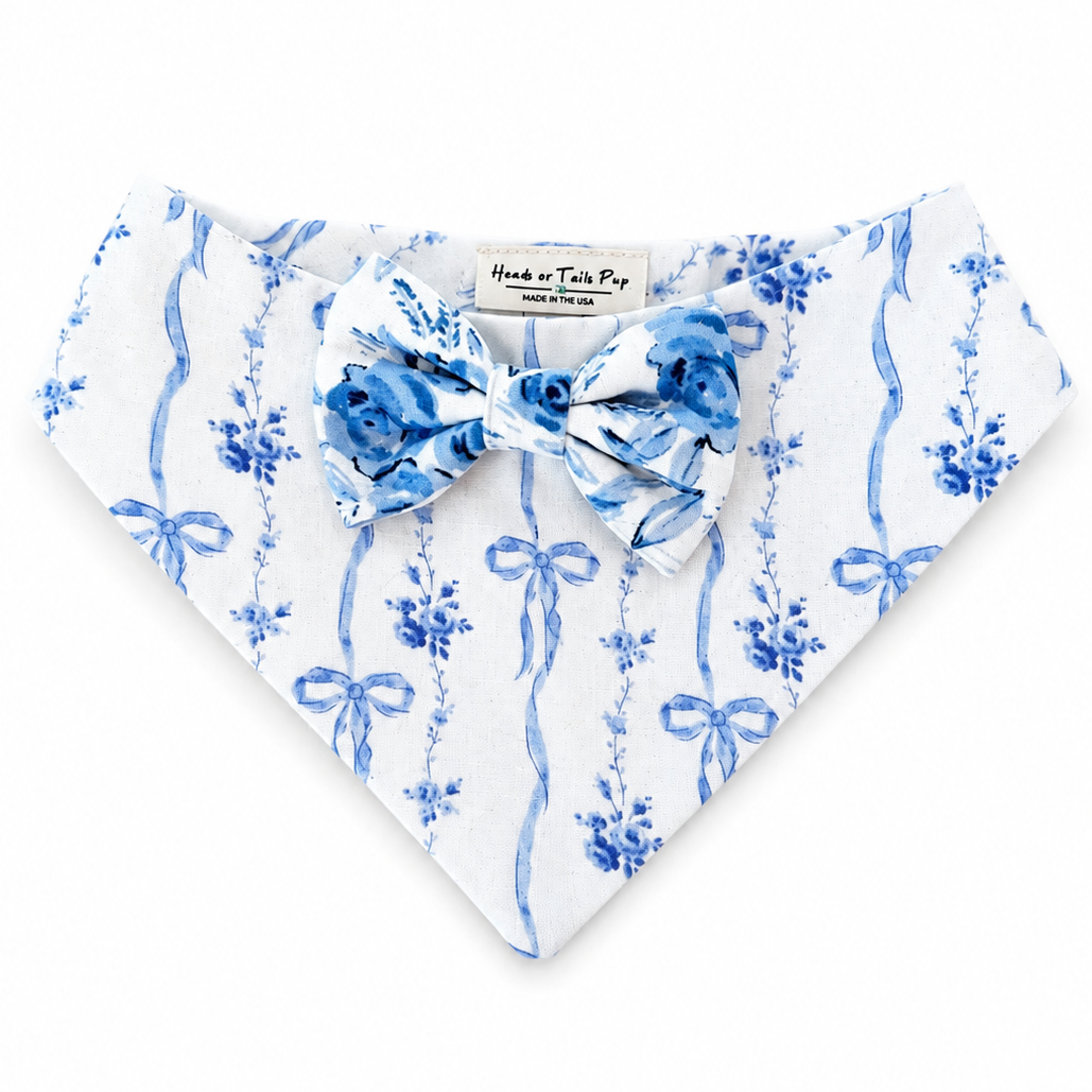 A white pet bandana with a blue floral and bow print, featuring a matching blue bow tie at the center, laid flat on a white background.