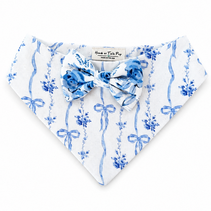 A white pet bandana with a blue floral and bow print, featuring a matching blue bow tie at the center, laid flat on a white background.