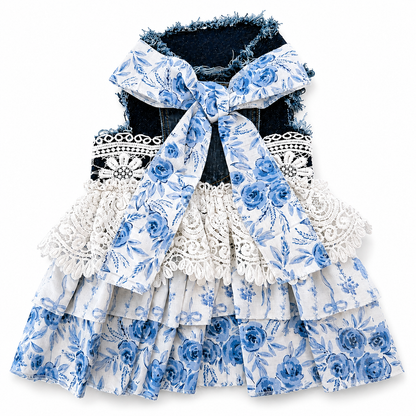 A sleeveless tiered dress for dogs, featuring a blue and white floral print, white lace ruffles, and a large matching fabric bow at the collar over a dark denim backing.