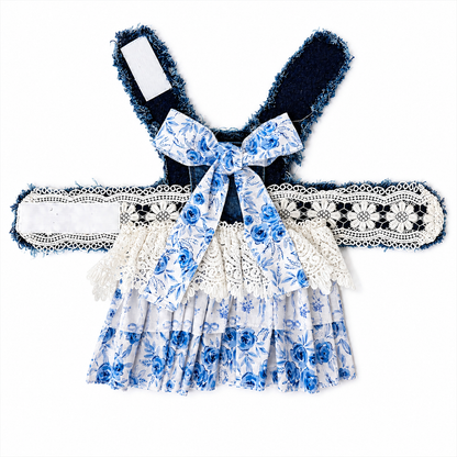 A pet dress with a blue and white floral print, featuring a ruffled skirt, a matching bow at the neckline, and wide lace-trimmed shoulder straps with dark blue fringed trim.