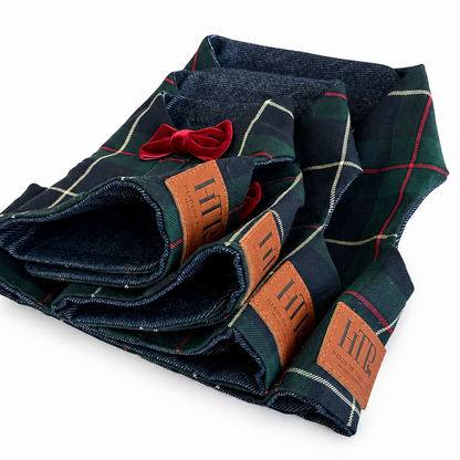 Folded plaid harnesses in dark navy and green with thin red and cream lines, featuring brown leather patches with 'HTP' branding and a small red bow, on a white background.