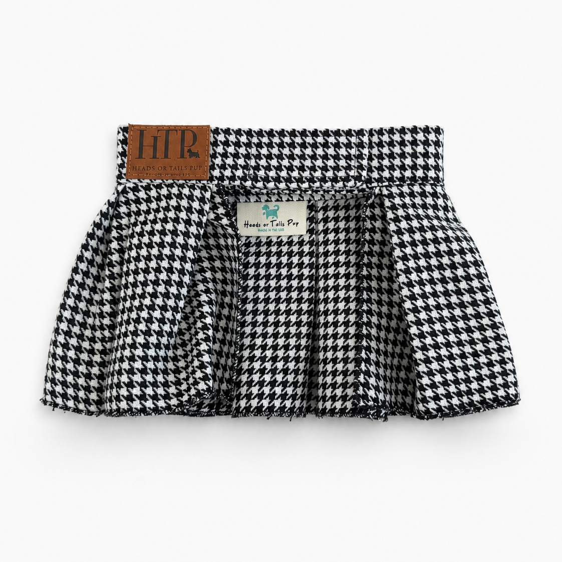 A black and white houndstooth pleated skirt with a brown leather brand patch on the waistband, photographed flat against a light gray background.