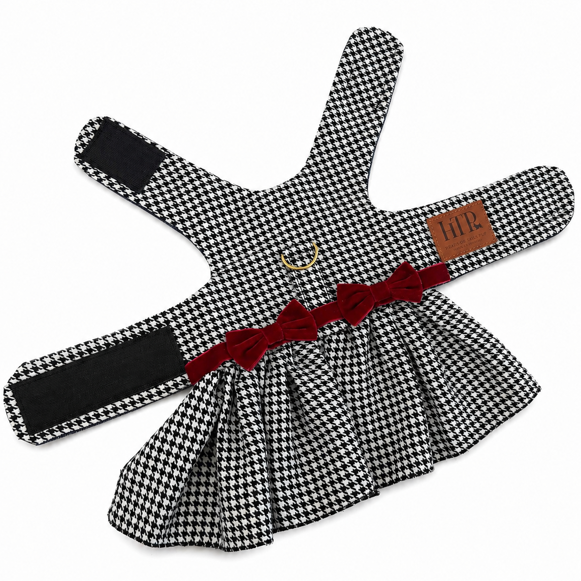 A black and white houndstooth pet harness with a red fabric bow at the center, a small gold metal ring, and a brown leather brand tag reading 'HTP'.