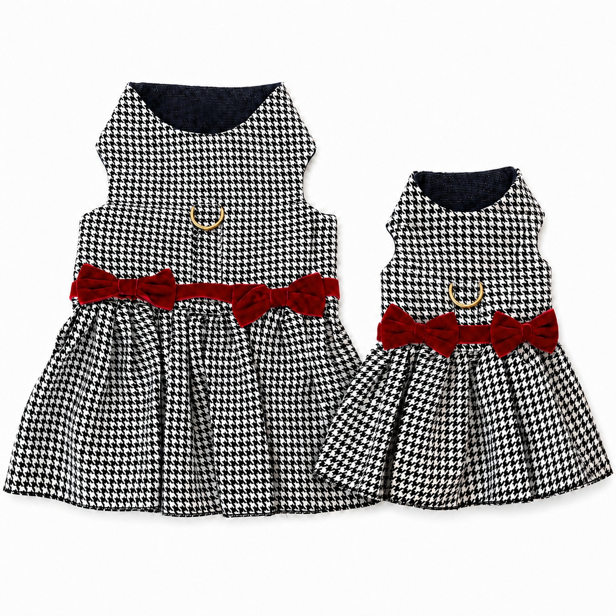 Two sleeveless houndstooth pet dresses shown on a plain white background, one larger dress and one smaller matching dress. Both have black-and-white small-scale check patterns, dark navy collars, red bow waist accents, and small gold D-rings.
