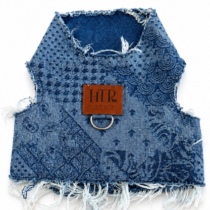A small blue patterned fabric vest or harness with frayed edges, a brown leather patch reading 'HTP', and a metal ring, photographed on a white background.