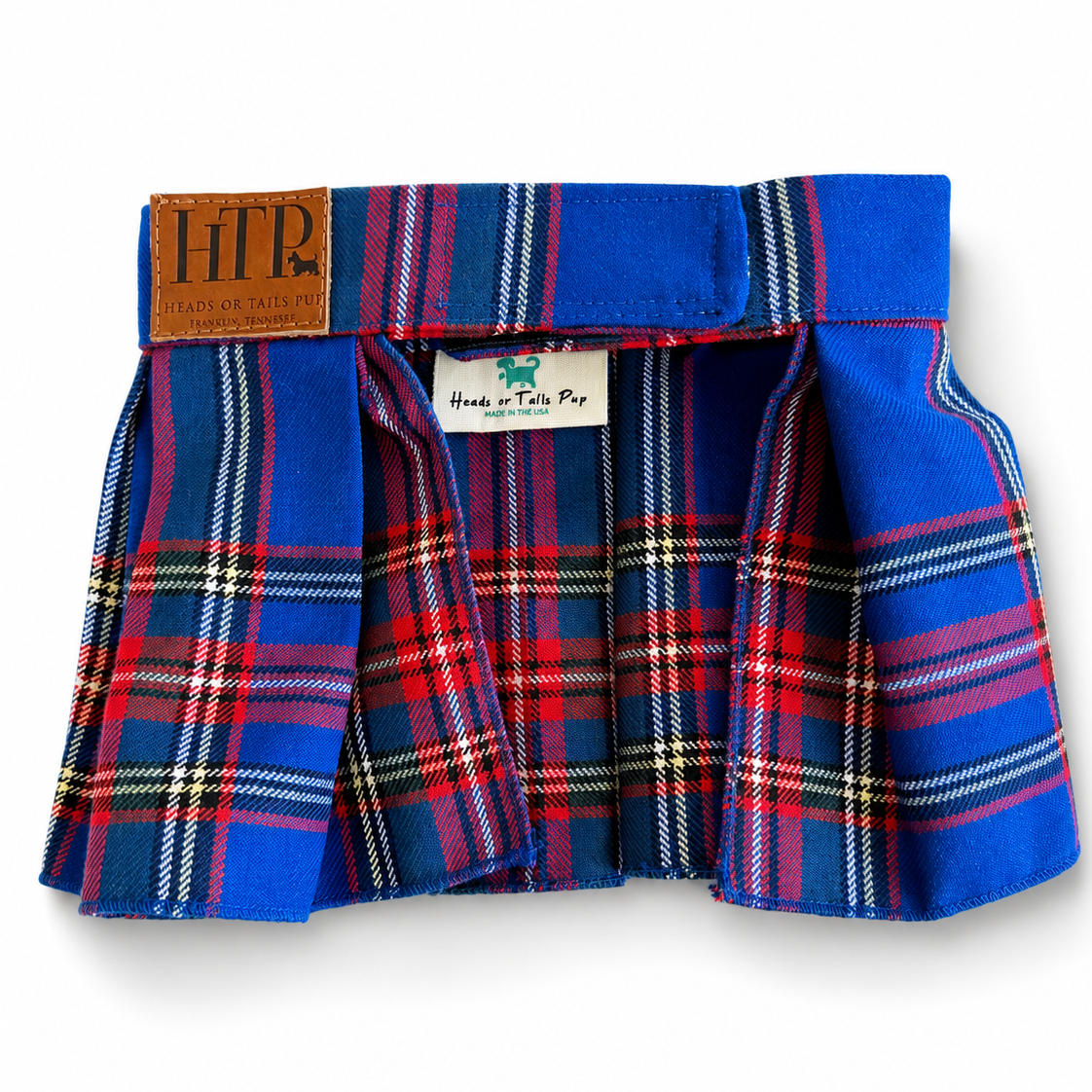 A small pleated tartan skirt in royal blue with red, white, and yellow plaid stripes, featuring a brown leather patch with 'HTP' on the left side and a white brand label in the center, shown on a white background.