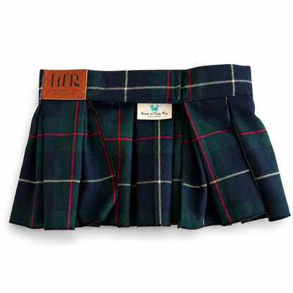 A pleated tartan mini skirt in navy blue with green, red, and white plaid stripes, featuring a brown leather brand patch on the left side and a white sewn-in label.