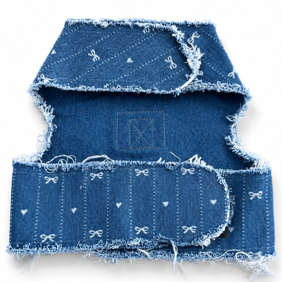 A folded denim harness with frayed edges and small white embroidered bow and heart motifs, shown on a plain white background.