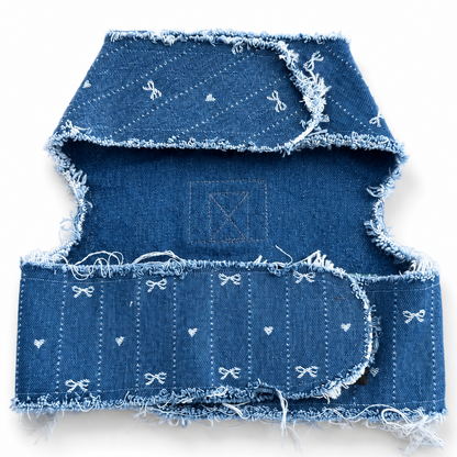 A folded denim harness with frayed edges and small white embroidered bow and heart motifs, shown on a plain white background.