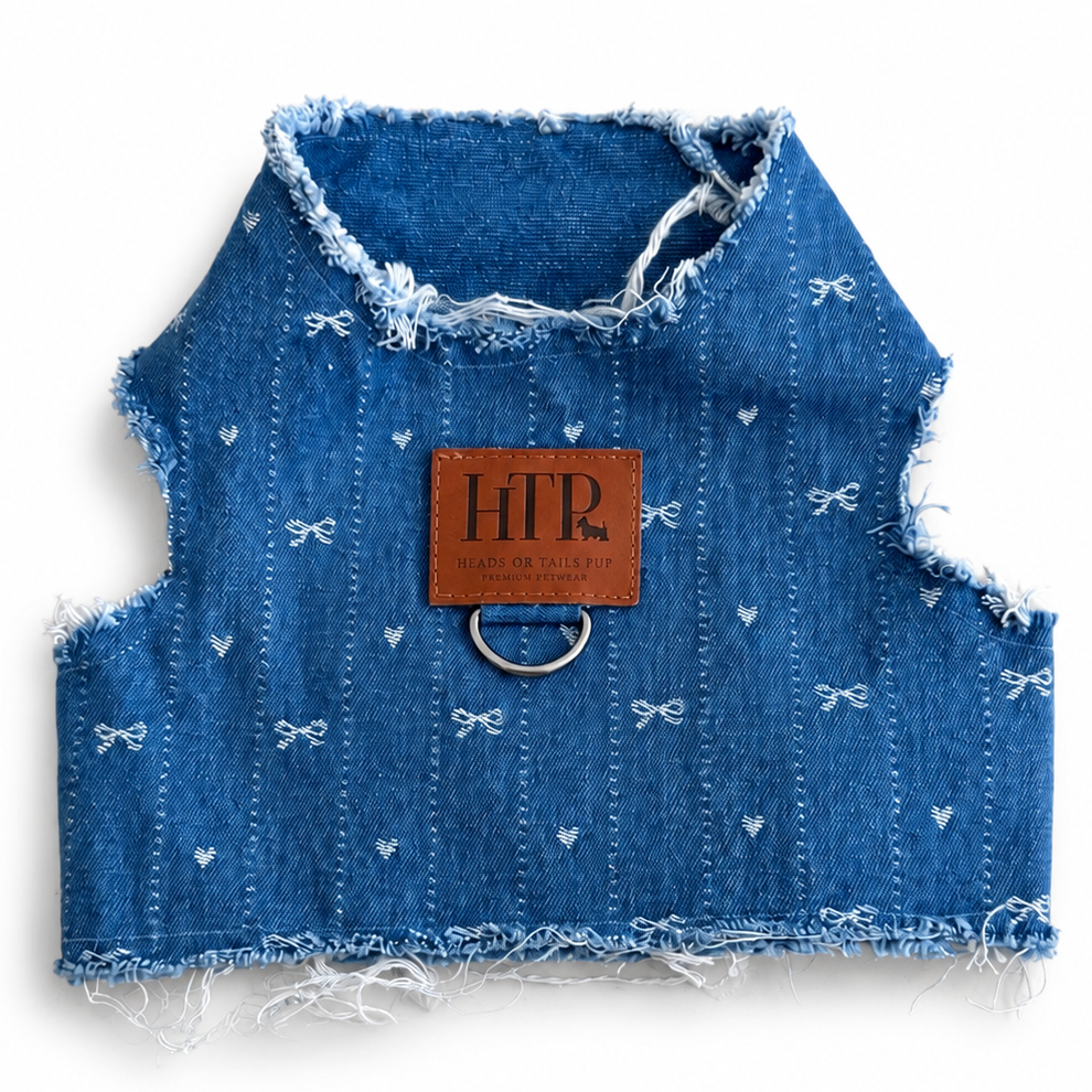 A blue denim-style pet harness with frayed edges, small white bow and heart motifs, and a brown leather brand patch reading 'HT' on a white background.