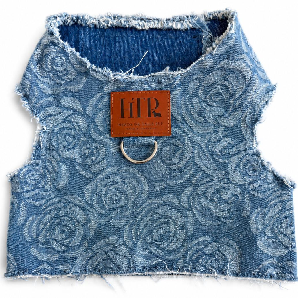 A blue denim pet harness with a tonal rose pattern, frayed edges, and a silver metal ring at the collar, featuring a brown leather brand patch that reads 'HTP' on a white background.