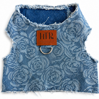 A blue denim pet harness with a tonal rose pattern, frayed edges, and a silver metal ring at the collar, featuring a brown leather brand patch that reads 'HTP' on a white background.